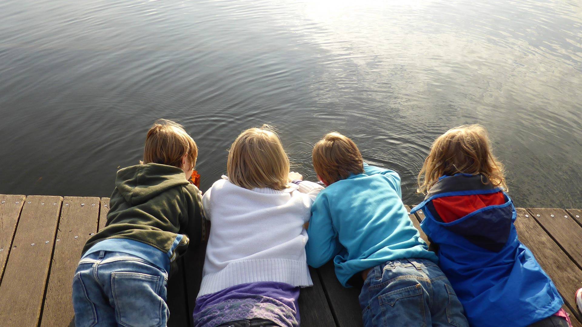 Quelle: pixabay.de / EME, https://pixabay.com/de/photos/kinder-m%C3%A4dchen-jungen-liegen-516340/ Vier Kinder liegen auf Steg und beobachten das Wasser